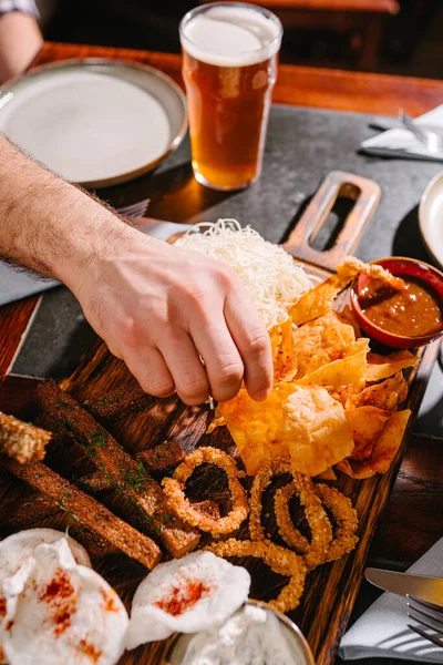 Tahta bir tahtanın üzerinde bira için abur cubur olan bir barda erkek eli. Patates ve pirinç cipsi, kızarmış ekmek, soğan halkaları, füme peynir, domates sosu, tartar sosu ve bira..