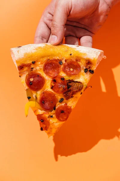 Piece of Pizza in hand with chorizo sausage, sweet peppers, sun dried tomatoes, dried chili peppers, gouda cheese and mozzarella cheese with pilati sauce, on a thin crust and on a fluffy board. orange background. copyright. top view