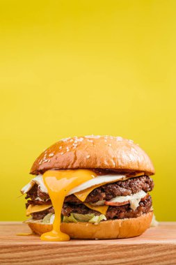 Çift köfteli burger, mermer biftek, pastırma, çedar peyniri, yumurta sarısı, domates, soğan, krema sosu ve ketçap. Tahta masa, sarı arka plan.