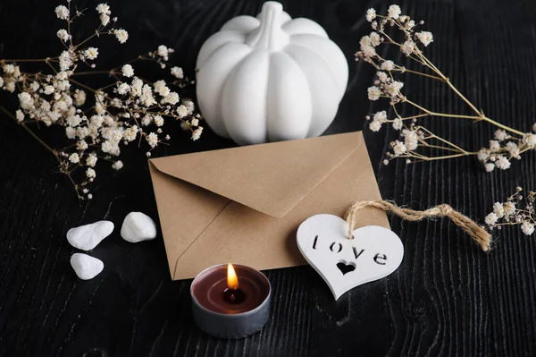 Mock-up card with Craft envelope, white heart and dry flowers on black wooden background. Still life with copyspace for text. Autumnal greeting card and advertisment