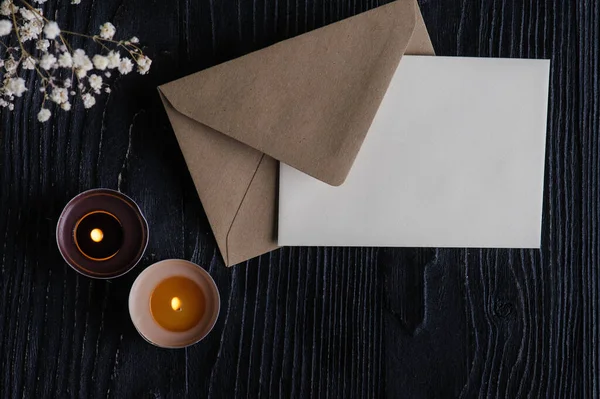 Mock-up card with Craft envelope, lit candles and dry flowers on black wooden background. Still life with copyspace for text. Autumnal greeting card and advertisment