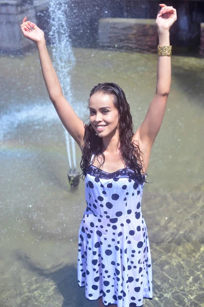 Glad tjej med upphöjda händer i en våt klänning och brunt hår poserar på bakgrund av fontänen — Stockfoto