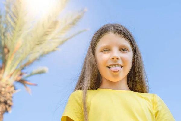A girl in a yellow T-shirt shows her tongue on the background of a palm tree in the summer.