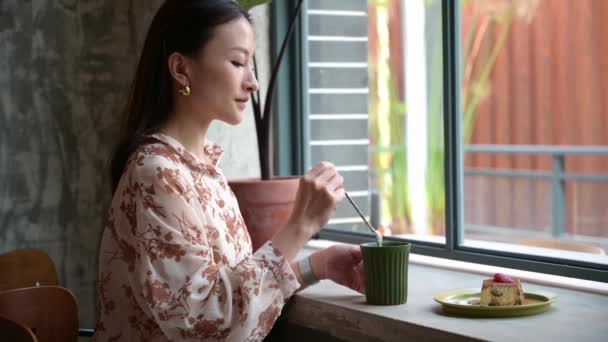 ガラスを身に着けているかなり中国系アジア人の若い女性は 窓の近くのカフェでラテを飲み それを見て コーヒー休憩中にリラックス 若い学生やビジネスマンがコーヒーショップで待っています — ストック動画