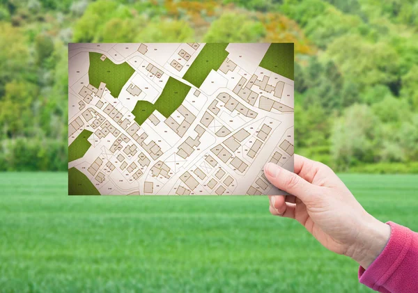 Land plot management - real estate concept with a vacant land on a green residential area available for building construction with hand holding a postcard about an imaginary cadastral map