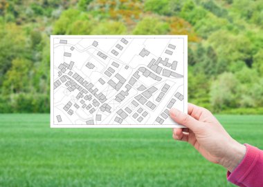 Arazi yönetimi - yeşil yerleşim alanındaki boş bir arazide, hayali bir kadastro haritasıyla ilgili bir kartpostalı elinde tutarak inşaat için kullanılabilir arazi kavramı
