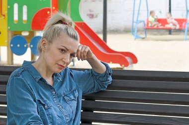Bir kadın kısırlıktan muzdarip, çocuk parkında üzgün oturuyor, arka planda oynayan çocukları seyrediyor. Kısırlık, kürtaj, doğum sonrası depresyon.