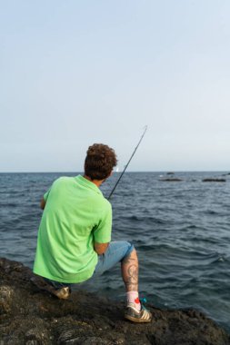 Balıkçı balıkçı kayalarda oturuyor, dönme tekniğini çalışıyor, arka planda kayalar ve deniz var.