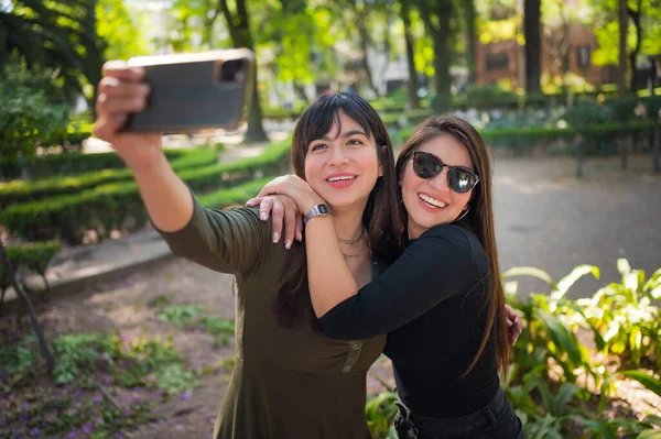 Two young latin female friends walking at the woods. Embrace each other and laughing. Friends making selfie. Two beautiful latin young women making selfie