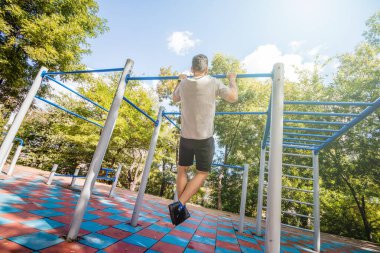  Güneşli bir günde, oyun parkında barfiks çeken bir adam..