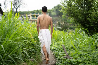 Young Indian fit boy, walking on a pathway beside crops in the field. An Indian priest walking while wearing white dhoti. Indian religious man.