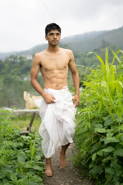 Young Indian fit boy, walking on a pathway beside crops in the field. An Indian priest walking while wearing white dhoti. Indian religious man.