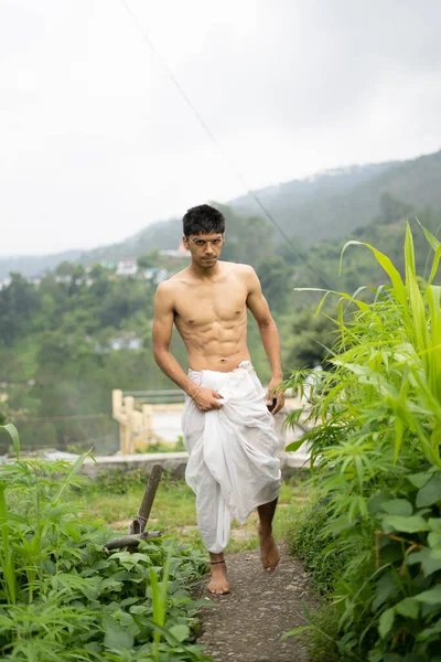 Young Indian fit boy, walking on a pathway beside crops in the field. An Indian priest walking while wearing white dhoti. Indian religious man.