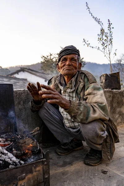 Almora, Uttarakhand - Jaunary 2 2022- An old man sitting in rugged clothes, soaking warmth from firewood. An village old man smiling into the camera.