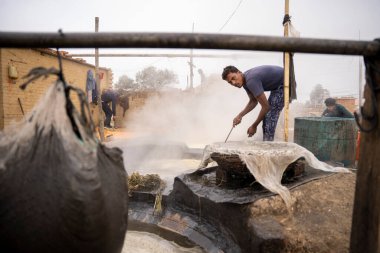 Garh Mukteshwar, Uttar Pradesh- 13 Ocak 2022 - UP 'deki Jaggery fabrikasında çalışan iki adam. Arka planda duman olan bir fabrikada çalışan insanlar..