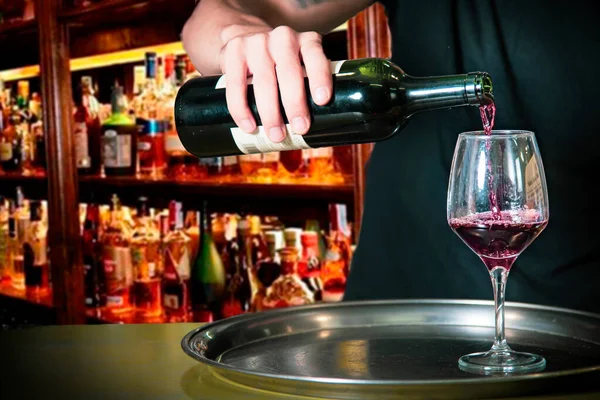 Barmen bardaki bir bardağa şarap koyuyor barın tezgahındaki tipik bir metal tepside, arka planda bir sürü alkol şişesi var.
