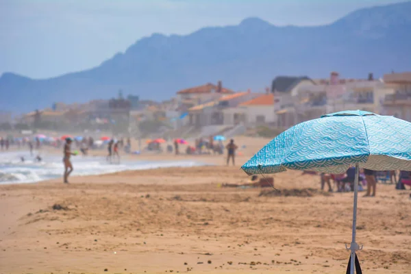 Fotoğrafın sağ tarafındaki mavi kumsal şemsiyesi, arka plandaki insanlar güneşin keyfini çıkarıyorlar.