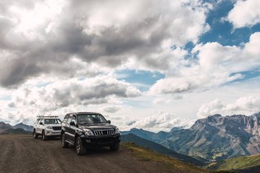 İki 4x4 off-road arabası, biri beyaz ve biri siyah, bulutlu bir günde arka planda dağlarla birlikte toprak bir pistte.