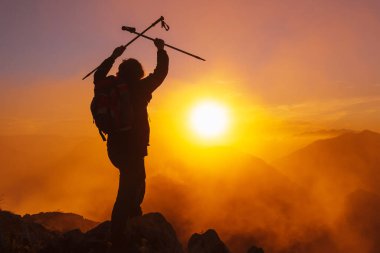 Sırt çantalı bir yürüyüşçü silueti zafer işareti olarak kollarını kaldırırken gün batımını izliyor. dağ sporu kavramı.