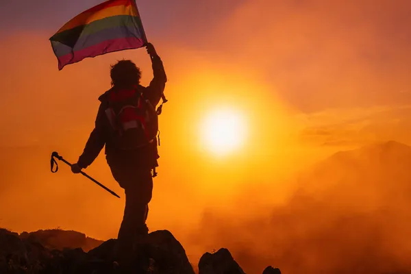 山の頂上で夕日を眺めながら虹のLgbtプライドの旗を背負ったバックパック付きハイカーのバックライト付きシルエット スポーツと健康的な生活の概念 — ストック写真