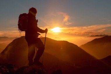 Sırt çantası ve sırıkla bir dağın tepesinde gün batımını seyreden bir yürüyüşçü silueti. spor ve sağlıklı yaşam konsepti.
