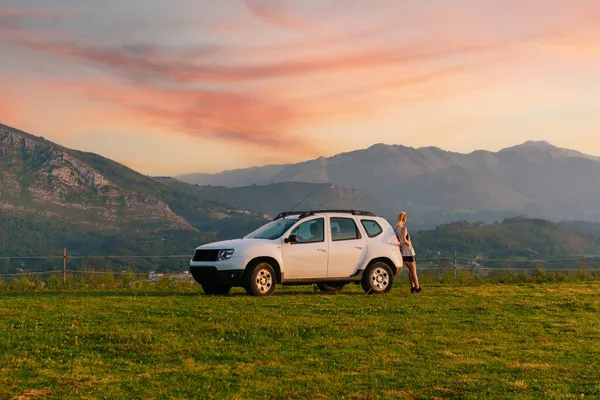 Beyaz 4x4 SUV 'una yaslanmış genç bir kadın macera gezisi sırasında gün batımını seyrediyor..