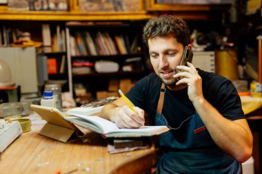 Atölyesindeki genç adam telefonla konuşuyor, siparişleri alıyor ve bir defter yazıyor..