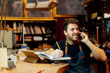 Atölyesindeki genç adam telefonla konuşuyor, siparişleri alıyor ve bir defter yazıyor..