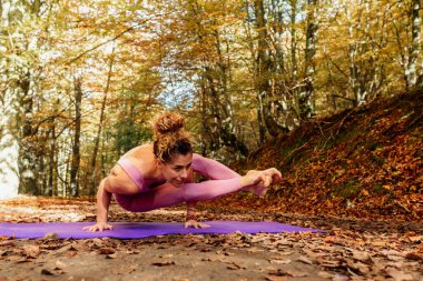 Yoga yapan güçlü bir kadın, Sekiz Açı egzersizi yapan, Astavakrasana pozu, asimetrik kol dengesi, sonbahar ormanlarında spor kıyafetleri giyen.