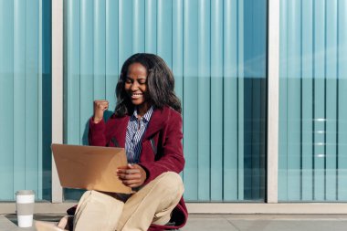 young african female student celebrating the success of her university exams. Black girl using a laptop sitting on the street.