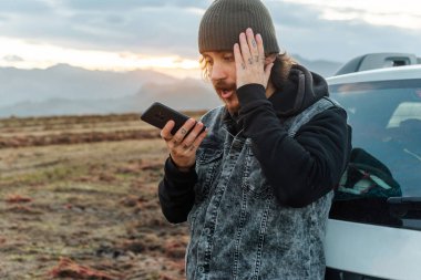 White middle-aged man with a beard and tattoos talking on the phone with the insurance company after a breakdown during a trip. Travel and adventure