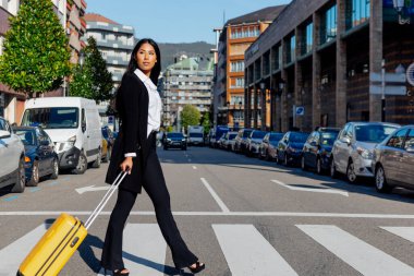 young latin business woman, elegantly dressed, crossing a crosswalk in the city with a yellow suitcase. business trip.