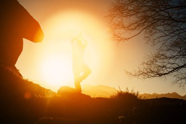 Yoga yapan bir adamın silueti, gün batımında dağdaki bir kayanın üzerinde ağaç pozu veren. sağlıklı yaşam tarzı