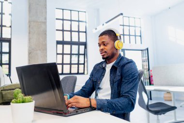 İş yerinde laptopla çalışan sarı kulaklıklı Afrikalı bir adam. Siyahi kolej çocuğu üniversite için okuyor ve çalışıyor..