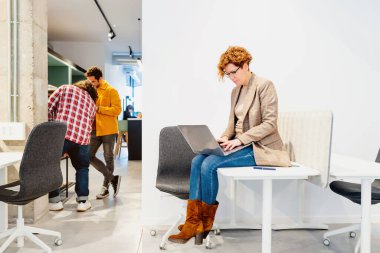 Orta yaşlı bir kadın, birlikte çalıştığı bir ofiste laptopuyla sıradan bir masada oturuyor..