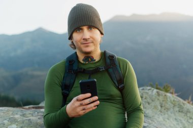 Orta yaşlı dağcı, yün şapkalı, sırt çantalı, piercingli ve dövmeli. Elinde cep telefonu olan kameraya bakıyor..