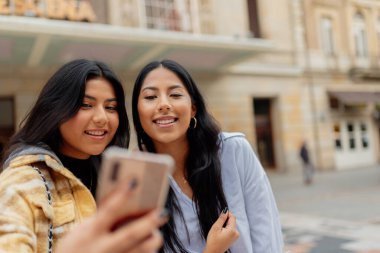 İki genç ve neşeli Latin kadın şehir turizmi sırasında cep telefonlarıyla fotoğraf çekiyor. Kız kardeşler birlikte iyi vakit geçiriyorlar.