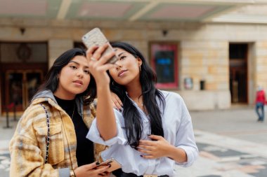 İki genç Latin kadın ve kız kardeş akıllı telefonlarıyla fotoğraf çekiyorlar. Kadınlar yolculuk sırasında sokakta selfie çekiyorlar..