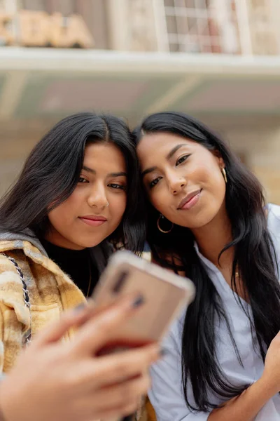 İki genç Latin kadın ve kız kardeş akıllı telefonlarıyla fotoğraf çekiyorlar. Kadınlar yolculuk sırasında sokakta selfie çekiyorlar..
