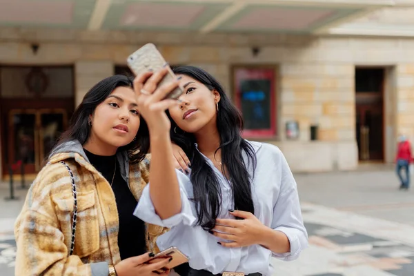 İki genç Latin kadın ve kız kardeş akıllı telefonlarıyla fotoğraf çekiyorlar. Kadınlar yolculuk sırasında sokakta selfie çekiyorlar..