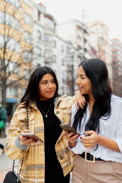 İki Latin kadın birbirine bakıyor ve sokakta cep telefonlarını tutuyorlar. İspanyol kız kardeş gezintide iyi vakit geçiriyor..