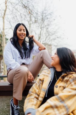 Şehir parkında oturmuş gülümseyen iki İspanyol kızın dikey portresi. İki kız kardeş birbirine rahatça bakar..