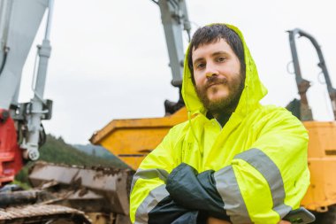 Kollarını kavuşturup kameraya bakan gülümseyen sakallı adam. Arka planda yansıtıcı yelek ve ağır makinelerle poz veren inşaat işçisi