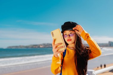 Genç Asyalı kadın şehir plajını gezerken cep telefonuyla fotoğrafını çekiyor. Çinli kız selfie çekiyor.