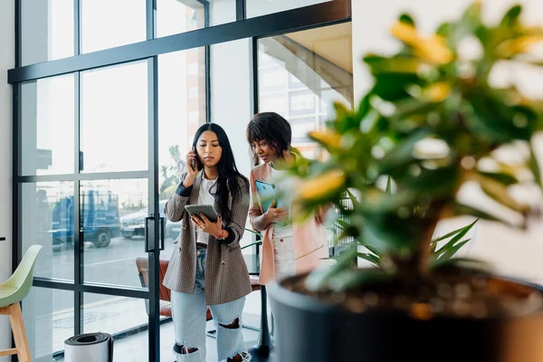 Two young business women of different races working in the office. Latin woman talks on the phone as she walks through the office with her co-worker.