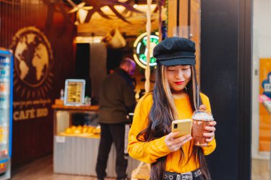 Asian woman using her mobile phone while drinking a special coffee with chocolate and cream in front of a modern coffee shop.