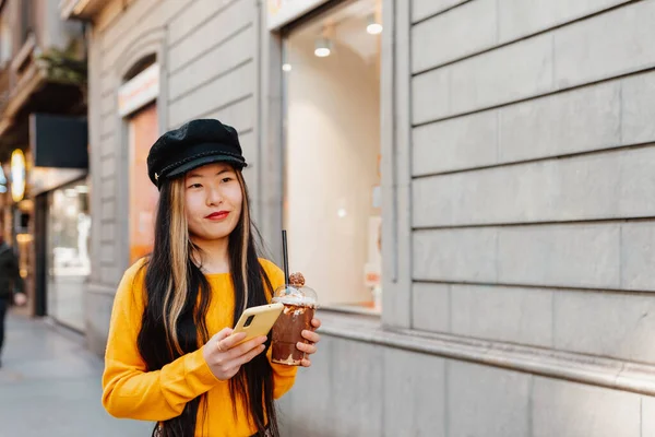 young asian woman, walking down a street in a european city while looking at her smartphone and having a chocolate milkshake. Smiling girl using map application on her phone while she is sightseeing.