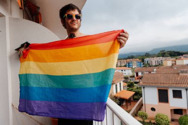 Evinin terasında gökkuşağı lgbt gurur bayrağı tutan dövmeli ve güneş gözlüklü bir adam. Gerçek insanlar gay gurur ayını kutluyor.