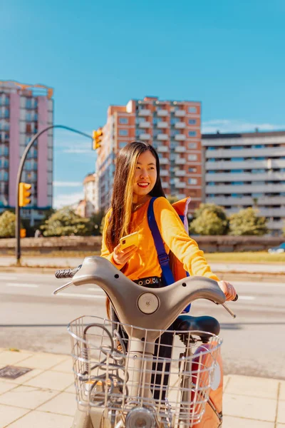 若いアジア系の女の子が携帯電話のアプリを使って都市移動のために自転車を借りています 持続可能な都市 — ストック写真
