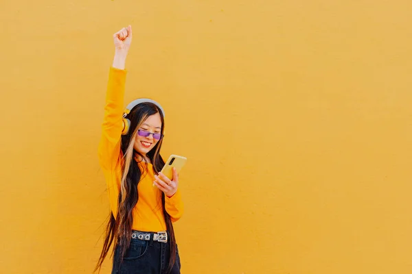 happy and smiling chinese woman listening to music with headphones from her smartphone with a yellow wall in the background.